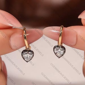 May include: A pair of gold hoop earrings, each featuring a heart-shaped pendant. The pendants have a clear, sparkling center stone surrounded by a dark border. The earrings are displayed against a soft background.