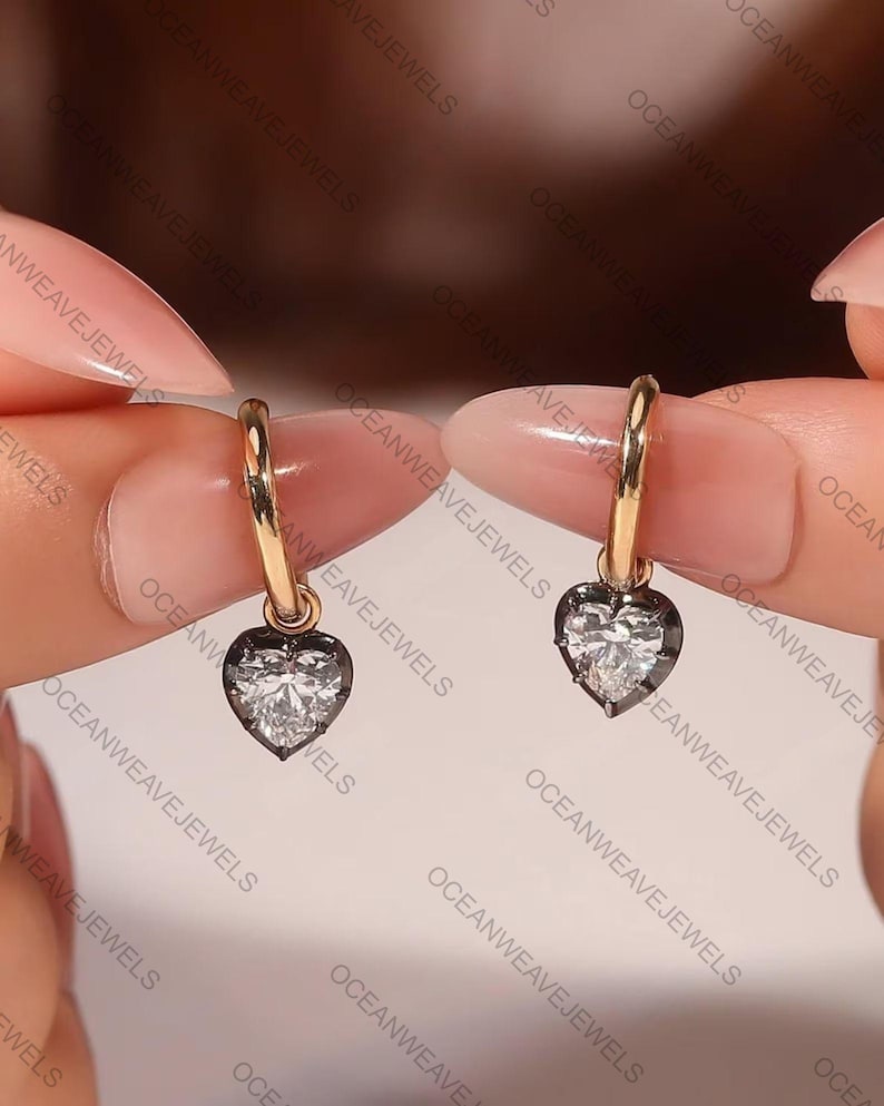 May include: A pair of gold hoop earrings, each featuring a heart-shaped pendant. The pendants have a clear, sparkling center stone surrounded by a dark border. The earrings are displayed against a soft background.