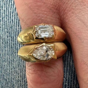 Peut inclure: Deux bagues en or avec de grosses pierres précieuses claires. La bague du haut présente une pierre rectangulaire taille émeraude, celle du bas une pierre en forme de poire. Les deux bagues ont une finition en or brossé et sont de style simple et élégant.