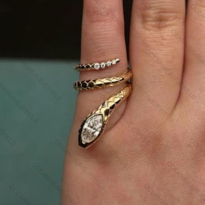 Könnte beinhalten: Ein goldfarbener Schlangenring mit strukturiertem Körper und einem diamantähnlichen Stein im Marquiseschliff am Kopf. Der Ring umschlingt den Finger, mit kleinen, runden Steinen am Schwanzteil. Ein einzigartiges Schmuckstück.