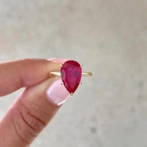 Könnte beinhalten: Ein goldener Ring mit einem großen, birnenförmigen, tiefroten Edelstein. Der Edelstein ist der Mittelpunkt, gefasst mit vier Krappen auf einem dünnen, goldenen Band. Der Ring wird vor einem neutralen Hintergrund präsentiert.