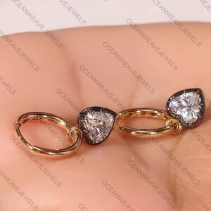 May include: A pair of gold hoop earrings with pear-shaped, clear gemstones set in a dark metal. The earrings are displayed against a neutral background. The brand name "OCEANWEAVEJEWELS" is visible.