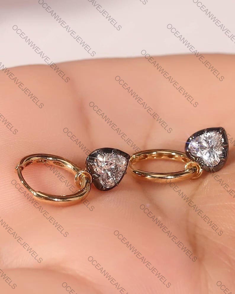 May include: A pair of gold hoop earrings with pear-shaped, clear gemstones set in a dark metal. The earrings are displayed against a neutral background. The brand name "OCEANWEAVEJEWELS" is visible.