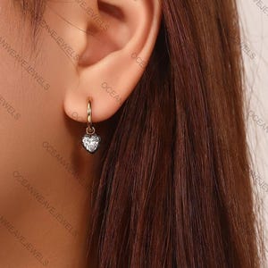 May include: A gold hoop earring with a heart-shaped, clear gemstone pendant. The earring is worn in an ear. The jewelry is from Ocean Weave Jewels.