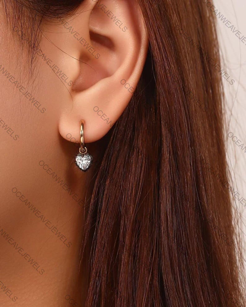 May include: A gold hoop earring with a heart-shaped, clear gemstone pendant. The earring is worn in an ear. The jewelry is from Ocean Weave Jewels.
