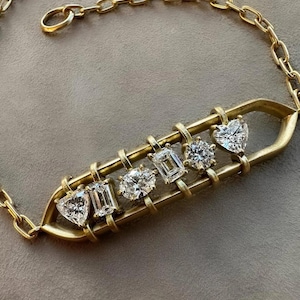 Puede incluir: Una pulsera dorada con una cadena y una barra central que sostiene varias piedras preciosas de diamantes. Los diamantes tienen varias formas, incluyendo corazón, esmeralda y talla redonda. La pulsera está colocada sobre un fondo neutro. La pulsera es de oro.