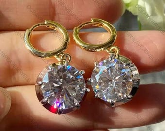 May include: Gold hoop earrings, each featuring a large, clear, round gemstone. The gemstones are cut to reflect light, creating a sparkling effect. The earrings are presented against a soft background, emphasising their brilliance.