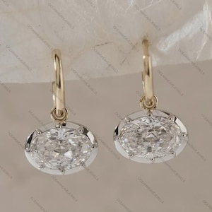 May include: A pair of gold hoop earrings with oval-shaped, clear gemstones. The gemstones are set in a white metal frame with a decorative edge. The earrings are displayed against a neutral background.
