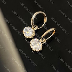 May include: A pair of gold hoop earrings, each featuring a dangling oval-cut clear gemstone. The stones are set in a prong setting, reflecting light with a brilliant sparkle. The metal has a polished gold finish, and the hoops have a hinged closure.