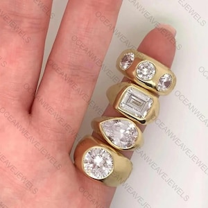 Könnte beinhalten: Eine Sammlung goldener Ringe mit verschiedenen Edelsteinformen, darunter rund, Smaragd und Tropfen. Die Ringe werden auf einem Finger präsentiert und zeigen ihre polierten Goldbänder und funkelnden Steine. Die Ringe sind von Oceanweave Jewels.