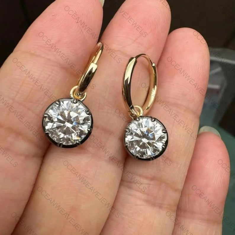 May include: A pair of gold hoop earrings with a large, round, clear gemstone pendant. The gemstones are set in a dark metal bezel. The earrings are displayed against a neutral background.