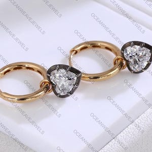 May include: A pair of gold hoop earrings with heart-shaped diamond pendants. The diamonds are set in a dark metal frame. The earrings are displayed on a white surface. The brand name "OCEANWEAVEJEWELS" is visible.