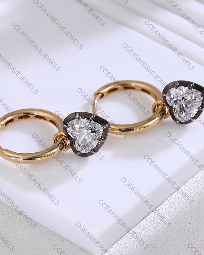 May include: A pair of gold hoop earrings with heart-shaped diamond pendants. The diamonds are set in a dark metal frame. The earrings are displayed on a white surface. The brand name "OCEANWEAVEJEWELS" is visible.