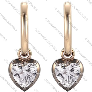 May include: A pair of gold-colored hoop earrings. Each earring features a heart-shaped, clear gemstone pendant. The gemstones are set in a dark metal frame. The earrings are displayed against a white background. The text "OCEANWEAVEJEWELS" is visible.