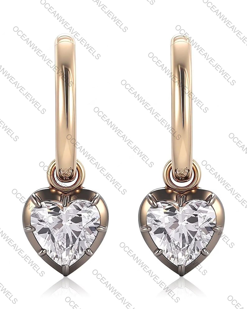May include: A pair of gold-colored hoop earrings. Each earring features a heart-shaped, clear gemstone pendant. The gemstones are set in a dark metal frame. The earrings are displayed against a white background. The text "OCEANWEAVEJEWELS" is visible.