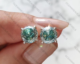 May include: A pair of stud earrings with large, round, faceted teal-green gemstones. The gemstones are set in silver-coloured metal. The earrings are shown against a light background.