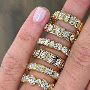 Könnte beinhalten: Eine Sammlung goldener Ringe mit verschiedenen Diamantschliffen, darunter Smaragd-, Rund- und Tropfenformen. Die Ringe sind in Goldbändern gefasst und an den Fingern einer Person präsentiert. Das Bild zeigt das Funkeln und Design der Ringe.
