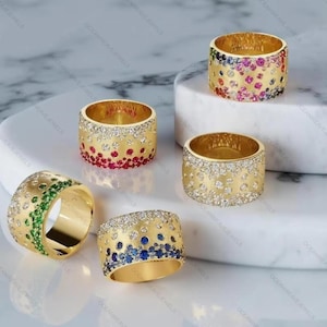 Puede incluir: Una colección de anillos de oro con diversas incrustaciones de piedras preciosas. Algunos anillos presentan un degradado de piedras rojas, rosas y azules, mientras que otros tienen piedras verdes o blancas. Los anillos se exhiben sobre una superficie blanca.