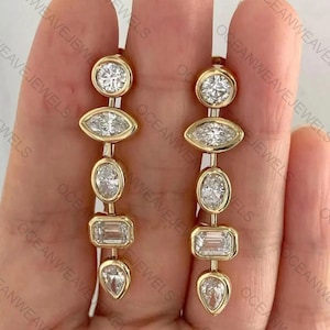 Pode incluir: Um par de brincos pendurados em ouro com diamantes redondos, marquesa, ovais, esmeralda e em forma de pêra. Os brincos são suspensos por uma fina corrente de ouro. O texto "OCEANWEAVEJEWELS" é visível. Uma joia de luxo.