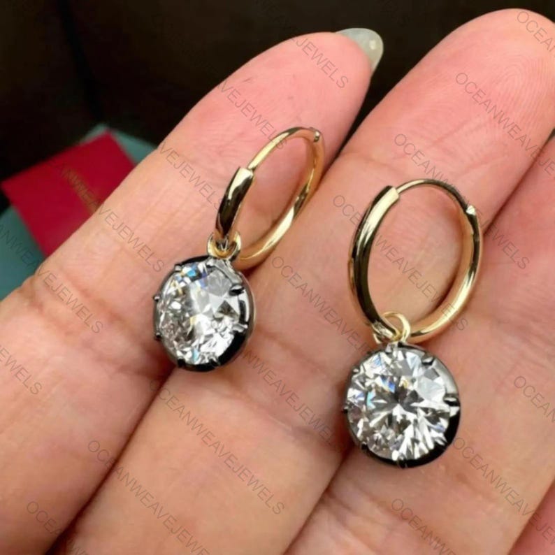May include: A pair of gold hoop earrings with a large, oval-cut, clear gemstone pendant. The gemstones are set in a dark metal bezel. The earrings are displayed against a neutral background.