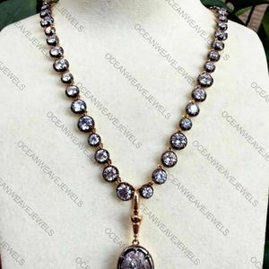 May include: A gold-toned necklace with a series of round, clear gemstones set in dark metal. The necklace has a pendant with an oval, clear gemstone. The text "OCEANWEAVEJEWELS" is visible in the image.