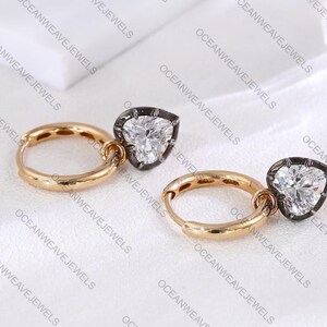 May include: A pair of gold hoop earrings with pear-shaped, clear gemstone pendants. The gemstones are set in a dark metal frame. The earrings are displayed on a white surface. The brand name "OCEANWEAVEJEWELS" is printed on the background.