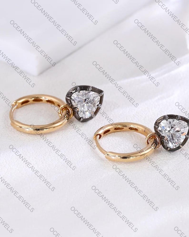 May include: A pair of gold hoop earrings with pear-shaped, clear gemstone pendants. The gemstones are set in a dark metal frame. The earrings are displayed on a white surface. The brand name "OCEANWEAVEJEWELS" is printed on the background.