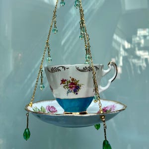 May include: A vintage teacup and saucer bird feeder. The white teacup has a blue base and floral design. The saucer and teacup are suspended by gold chains with green and blue beads. A unique garden decor item.