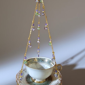 May include: A white porcelain teacup and saucer with gold trim, suspended by three gold-toned metal chains. The chains are adorned with small, multi-colored purple and green faceted beads, converging at a single point above the teacup.