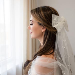 Peut inclure: Profil d'une personne portant un voile de tulle blanc orné d'un grand nœud et de perles. Le voile est fixé dans les cheveux. La personne regarde vers la gauche, avec un arrière-plan doux et neutre.