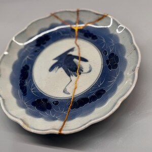 Könnte beinhalten: Runder Teller aus weißer und blauer Keramik mit Kranich-Illustration. Der Teller hat einen gewellten Rand und ist mit goldfarbenem Kintsugi repariert. Dekorativer Artikel.