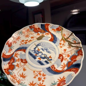 May include: A white ceramic plate with a scalloped edge, featuring a central blue and white design and red and orange floral patterns. The plate has been repaired with gold accents, showcasing the Japanese art of Kintsugi.