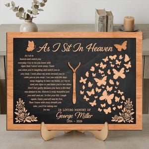 Pode incluir: Placa memorial emoldurada em madeira com o texto "As I Sit In Heaven" e um poema. A placa apresenta borboletas e detalhes florais cortados a laser. O memorial é dedicado a George Miller, com os anos 1954-2026.