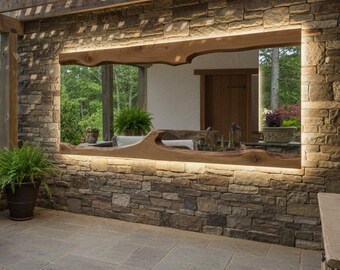 May include: Outdoor patio with a stone wall and a wooden pergola. A large, rustic wooden frame with integrated lighting surrounds a view of the outside. A wicker chair with a cream cushion sits on the patio.