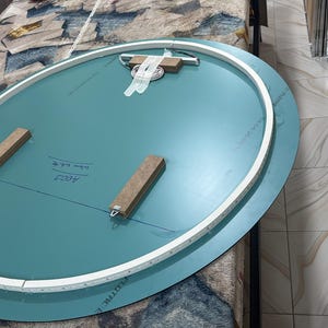 Peut inclure: Un dos de miroir ovale pr&eacute;sentant une surface bleu sarcelle avec un bord de cadre blanc. Deux blocs de bois rectangulaires sont fix&eacute;s au centre, et un support de suspension en m&eacute;tal avec du ruban adh&eacute;sif blanc est plac&eacute; sur le bord sup&eacute;rieur.