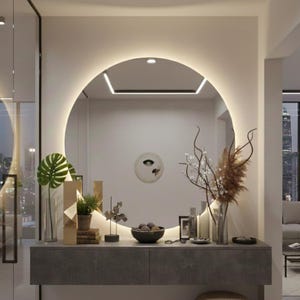 Peut inclure: Un grand miroir mural circulaire avec r&eacute;tro&eacute;clairage LED int&eacute;gr&eacute;, fix&eacute; au-dessus d'une console flottante gris fonc&eacute;. Le miroir refl&egrave;te une pi&egrave;ce avec une d&eacute;coration murale ronde, tandis que la console supporte une plante verte, des objets d&eacute;coratifs et des tiges florales s&eacute;ch&eacute;es dans un vase en verre.