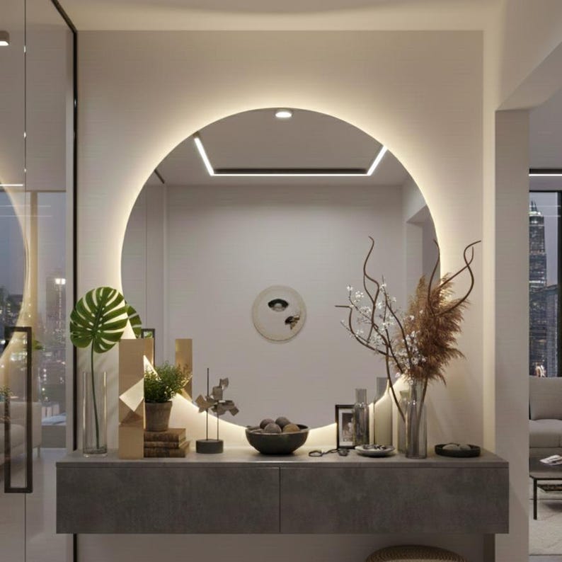 Peut inclure: Un grand miroir mural circulaire avec r&eacute;tro&eacute;clairage LED int&eacute;gr&eacute;, fix&eacute; au-dessus d'une console flottante gris fonc&eacute;. Le miroir refl&egrave;te une pi&egrave;ce avec une d&eacute;coration murale ronde, tandis que la console supporte une plante verte, des objets d&eacute;coratifs et des tiges florales s&eacute;ch&eacute;es dans un vase en verre.