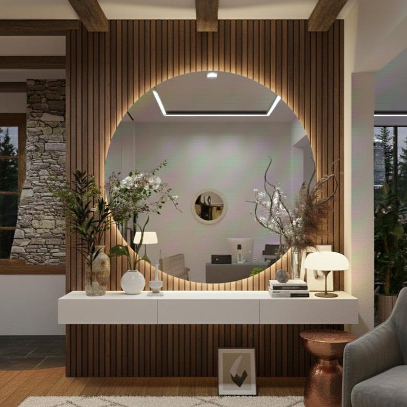Peut inclure: Un grand miroir mural circulaire avec r&eacute;tro&eacute;clairage LED int&eacute;gr&eacute;, fix&eacute; sur un mur &agrave; lattes de bois verticales au-dessus d'une longue console flottante blanche. La console supporte deux vases en verre avec des fleurs, des livres et une lampe.