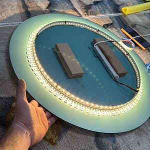 Peut inclure: Un miroir circulaire dot&eacute; d'une bande lumineuse LED int&eacute;gr&eacute;e dispos&eacute;e en anneau sur sa surface. Deux blocs de bois rectangulaires sont fix&eacute;s au centre, et un petit composant &eacute;lectronique est reli&eacute; &agrave; la bande lumineuse par des fils.