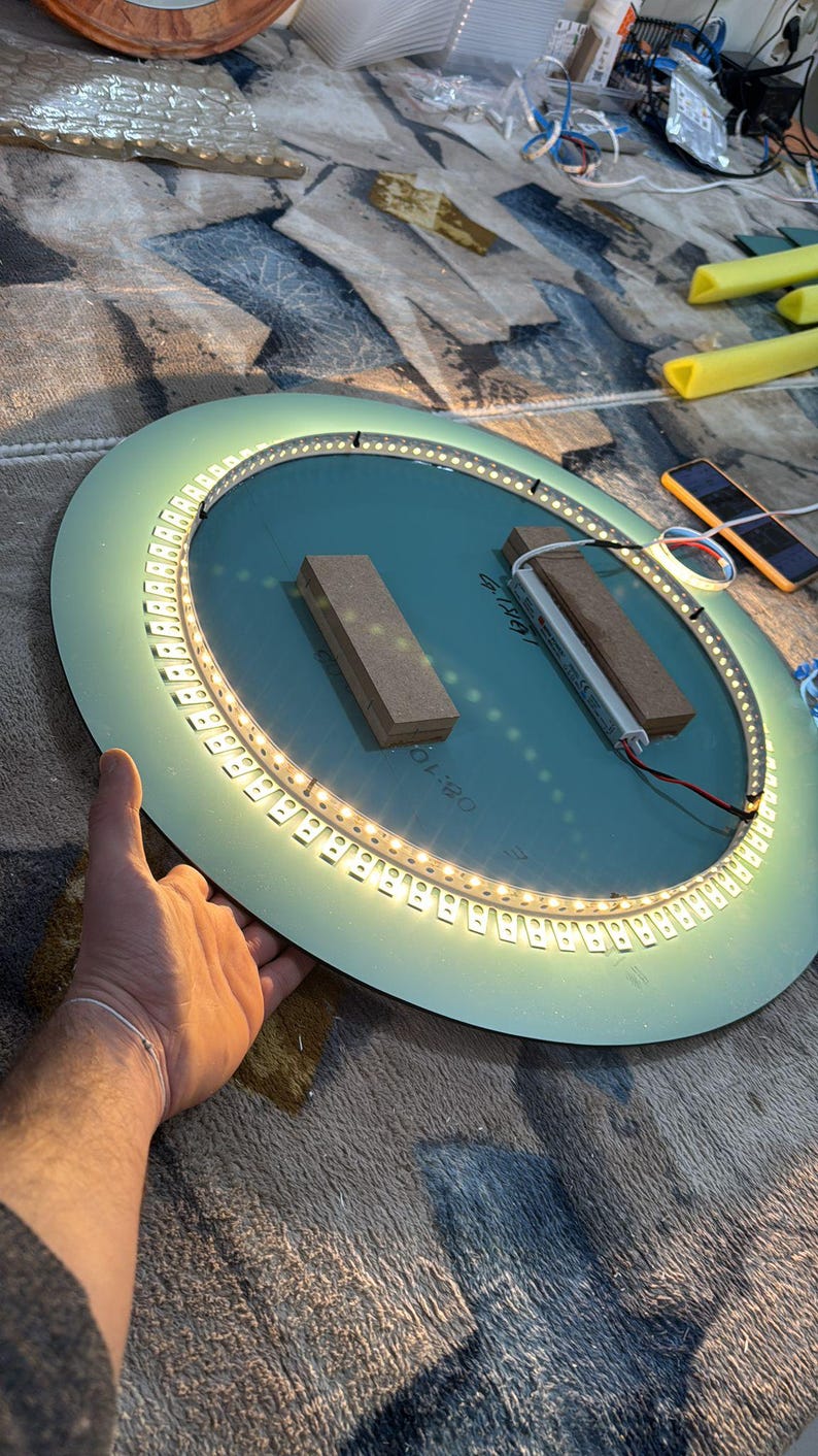 Peut inclure: Un miroir circulaire dot&eacute; d'une bande lumineuse LED int&eacute;gr&eacute;e dispos&eacute;e en anneau sur sa surface. Deux blocs de bois rectangulaires sont fix&eacute;s au centre, et un petit composant &eacute;lectronique est reli&eacute; &agrave; la bande lumineuse par des fils.