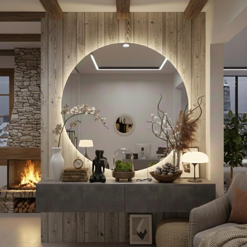 Peut inclure: Un grand miroir mural circulaire avec r&eacute;tro&eacute;clairage LED int&eacute;gr&eacute; mont&eacute; sur un mur en panneaux de bois verticaux. Le miroir refl&egrave;te l'int&eacute;rieur d'une pi&egrave;ce, tandis qu'une console grise en dessous porte un vase blanc, des livres et divers objets.