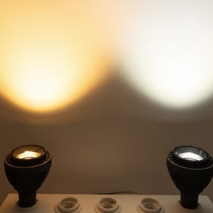 Peut inclure: Deux projecteurs LED noirs mont&eacute;s sur une base blanche. Le projecteur de gauche &eacute;met une lumi&egrave;re jaune chaude, tandis que celui de droite &eacute;met une lumi&egrave;re blanche froide. Les deux projettent des faisceaux circulaires sur le mur.