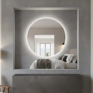Peut inclure: Un grand miroir mural circulaire avec r&eacute;tro&eacute;clairage LED int&eacute;gr&eacute;, mont&eacute; dans une alc&ocirc;ve rectangulaire encastr&eacute;e. Le miroir refl&egrave;te l'int&eacute;rieur d'une chambre avec un lit aux draps blancs, des oreillers gris et un jet&eacute; de lit gris textur&eacute;.