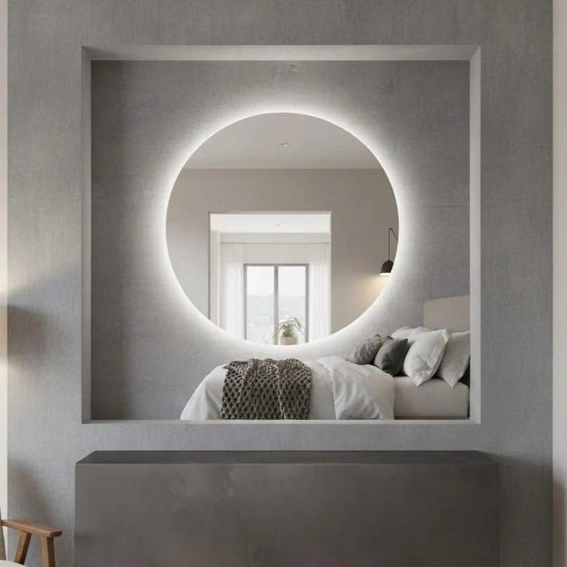 Peut inclure: Un grand miroir mural circulaire avec r&eacute;tro&eacute;clairage LED int&eacute;gr&eacute;, mont&eacute; dans une alc&ocirc;ve rectangulaire encastr&eacute;e. Le miroir refl&egrave;te l'int&eacute;rieur d'une chambre avec un lit aux draps blancs, des oreillers gris et un jet&eacute; de lit gris textur&eacute;.