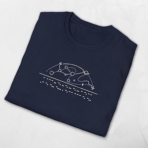 Könnte beinhalten: Marineblaues T-Shirt mit weißer Grafik einer Sternenkonstellation mit gepunkteten Linien und Sternen. Unterhalb der Konstellation befindet sich eine Reihe von Punkten. Das T-Shirt ist gefaltet.