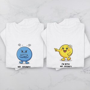 Puede incluir: Dos camisetas blancas con gráficos de dibujos animados. Una presenta un personaje azul llamado "MR. GRUMPY" con una expresión enfadada. La otra muestra un personaje amarillo con una flor, señalando, y el texto "I'M WITH MR. GRUMPY."