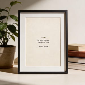 May include: A black rectangular picture frame containing a cream-colored print with the text "do small things with great love." and "- mother teresa" in a typewriter-style font, centered above a small decorative leaf illustration.