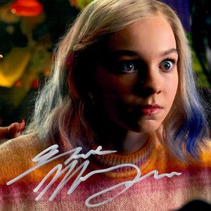 May include: A person with blonde hair and blue eyes, wearing a colorful sweater with a signature. The person's hand is extended with long, painted nails. A red watch is visible on the wrist.