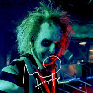 Może przedstawiać: Zbliżenie postaci z filmu Beetlejuice, z zielonymi włosami i czarno-białą koszulą w paski. Twarz postaci jest pomalowana zielonym i czarnym makijażem. Na koszuli widoczny jest podpis.