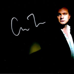 May include: A portrait of a person in a dark suit and white shirt, with a handwritten signature in white. The person is looking directly at the camera. The background is black, with a hint of yellow light.